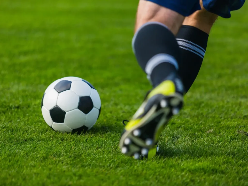 Gros plan sur la jambe d'un joueur de socca portant des crampons jaunes et noirs, frappant puissamment le ballon sur la pelouse verte du terrain.