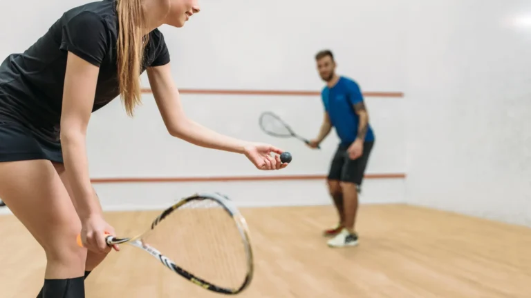 Une femme en tenue de sport sert une balle de squash sur un court intérieur. À l'arrière-plan, un homme en chemise bleue s'apprête à frapper la balle, raquette à la main.