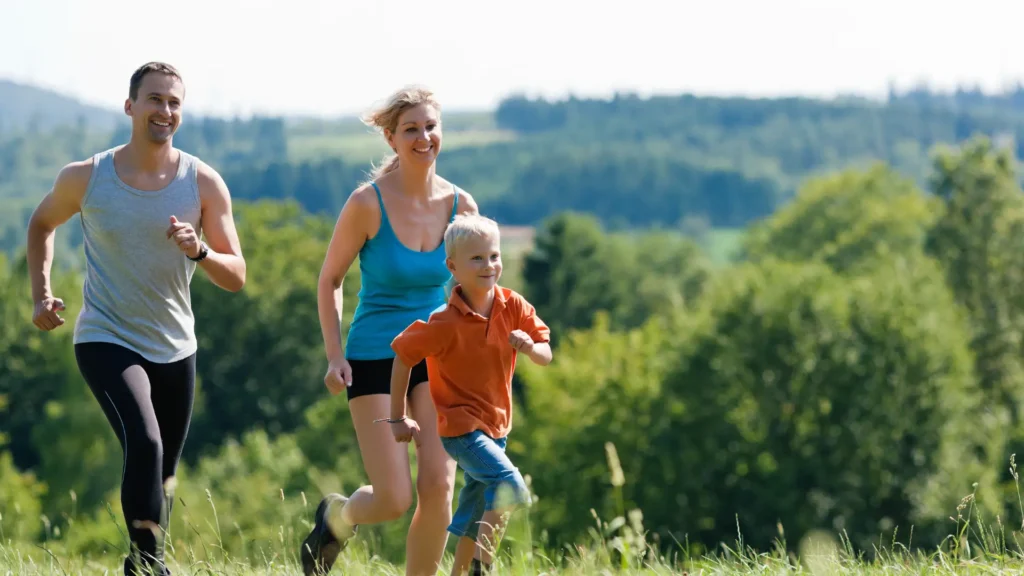 Une famille — un homme en t-shirt gris, une femme en haut bleu et un petit garçon en t-shirt orange — court joyeusement ensemble à travers une prairie verdoyante dans les montagnes, avec une forêt et des collines en arrière-plan. Tous sourient.
