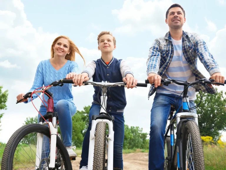 Une famille — une femme, un garçon et un homme — roule côte à côte à VTT sur un chemin forestier par une journée ensoleillée. Ils regardent tous l'objectif et semblent très heureux.