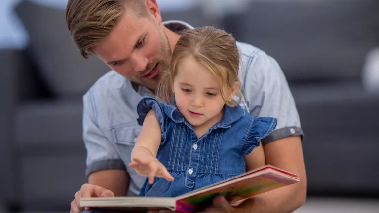 Mężczyzna z krótkimi blond włosami i brodą siedzi na kanapie, trzymając na kolanach małą dziewczynkę w niebieskiej sukience. Oboje z zainteresowaniem patrzą w otwartą książkę z ilustracjami, którą trzymają razem. Dziewczynka wskazuje palcem na stronę książki, a mężczyzna uśmiecha się ciepło.
