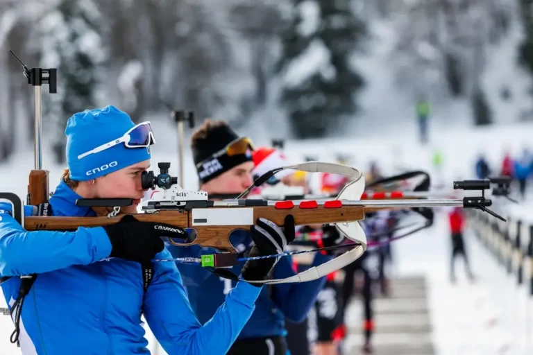 Des concurrents en biathlon tirent sur des cibles avec des fusils lors d'une compétition hivernale, avec une piste enneigée et d'autres athlètes en arrière-plan.
