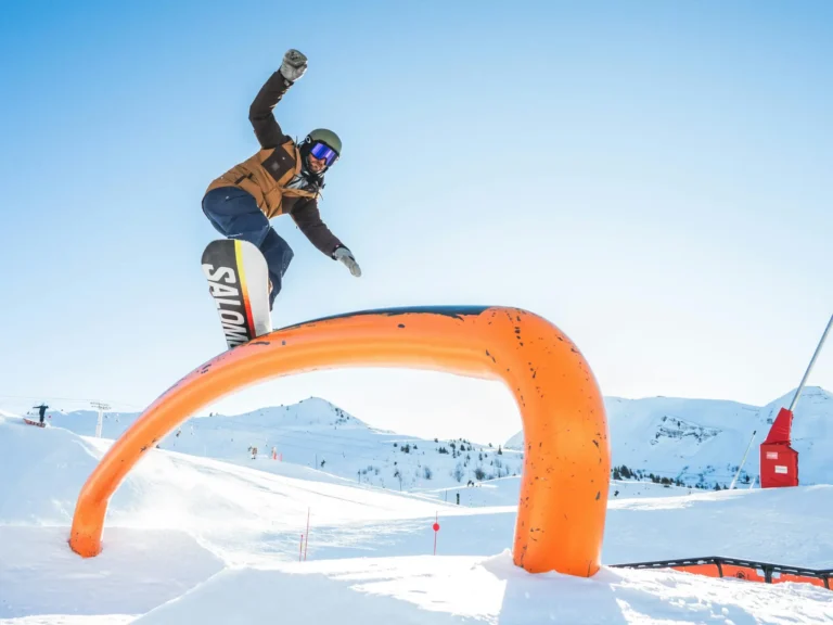 Un snowboardeur effectue un saut sur un rail orange, un saut en hauteur avec la main levée, des montagnes enneigées en arrière-plan