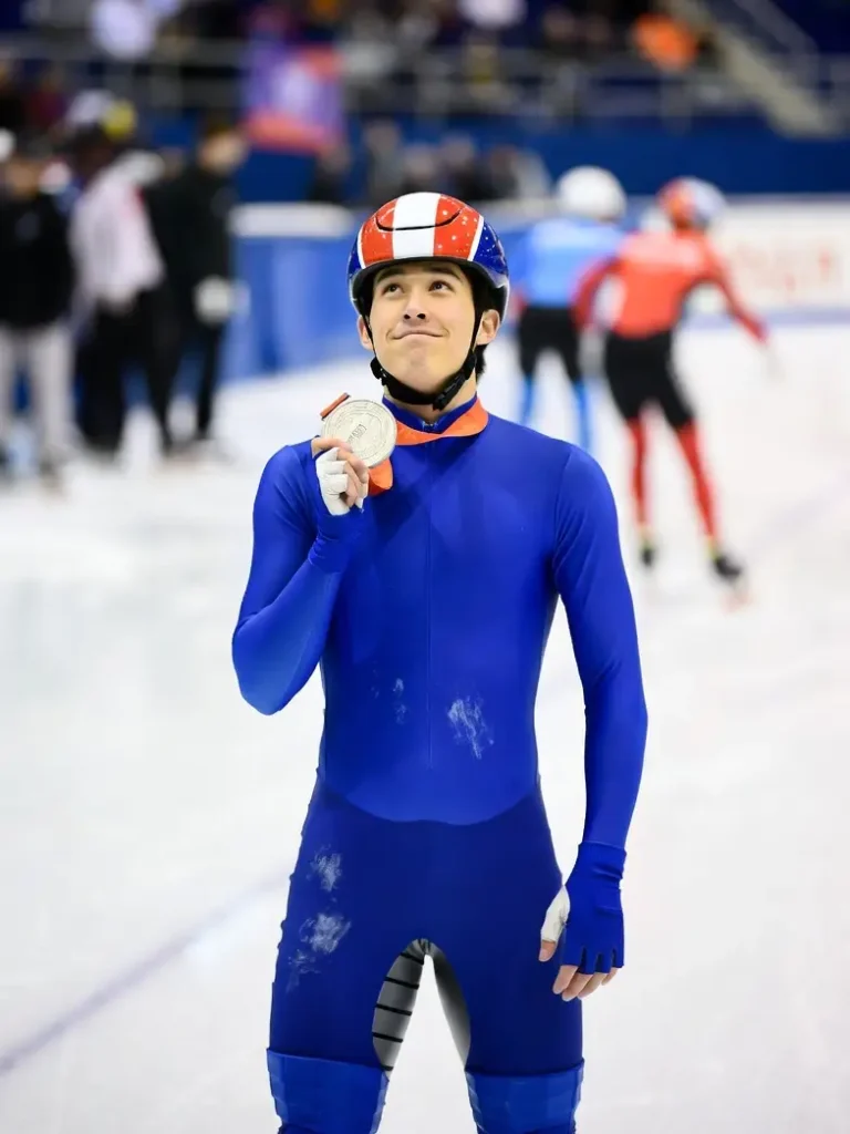 Un patineur de short-track après sa course – un jeune athlète en combinaison et casque bleu, rouge et blanc, tenant une médaille d'argent olympique. Il esquisse un sourire, la tête levée, debout sur la patinoire, entouré des tribunes du palais olympique.